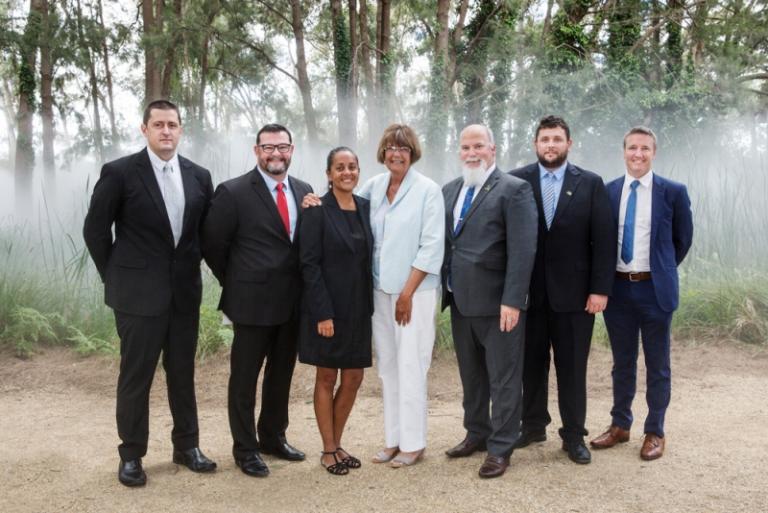 Men and women in business attire stand in a line in front of trees.