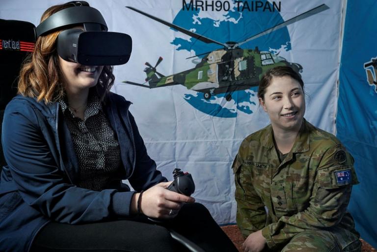 A woman in an Australian Army camouflage uniform is kneeling in front of a white and blue sheeted background with a picture of a MRH90 Taipan helicopter. She is next to a woman in business attire with a virtual reality headset on, who is sitting on a heli