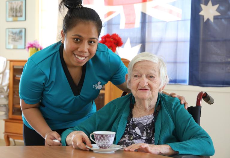 Photograph of an older woman having a cup of tea, posing with a younger female care worker.