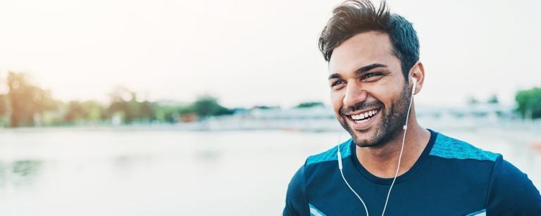 Picture of a male presenting human wearing headphones while outside. He has a smile on his face.