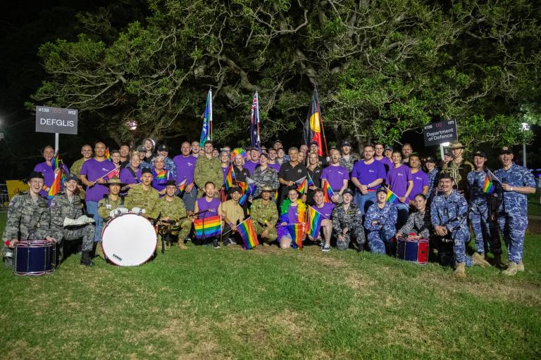 Colourful and diverse group from the Australian Defence Force and Defence Civilian cohort in Hyde Park, Sydney. One person a DEFGLIS sign, and another holds an Australian Defence Force sign, the Torres Strait Islander flag, Australian flag and Australian Aboriginal flag are shown in the background. Some members of the group are holding rainbow flags, and some are seated on the group with musical instruments.