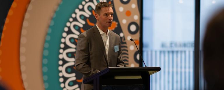 Image of Gary Rake standing at a lectern presenting to a crowd. Behind him is Aboriginal artwork.