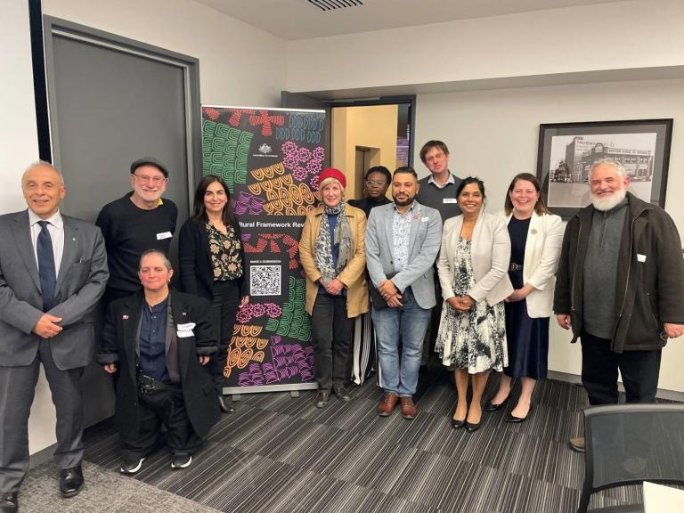 Group of attendees at a Multicultural Framework Review meeting standing in front of a colourful banner smiling at the camera.