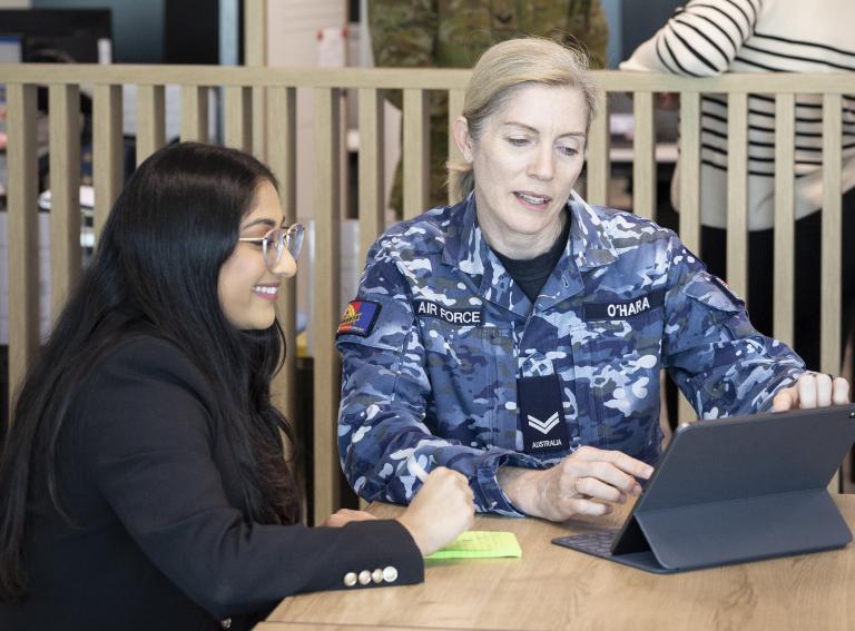 An Airforce staff member in uniform and non ADF staff member in business casual attire sitting at a table both looking at a digital tablet. The ADF staff member is scrolling on the tablet screen while other staff member is smiling.