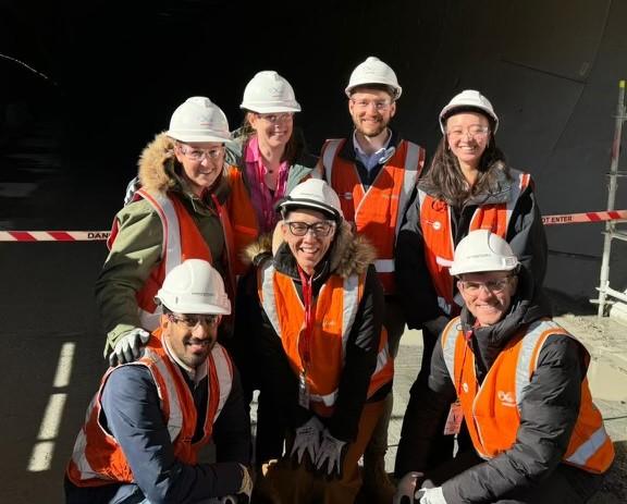 The Finance Snowy Hydro Shareholder team wearing safety glasses, orange high-visibility vests and white hard hats grouped together at the Snowy 2.0 construction site, smiling and looking at the camera. 