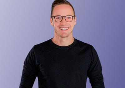 Picture of a person in glasses with short dark hair and a black top standing in front of a purple background