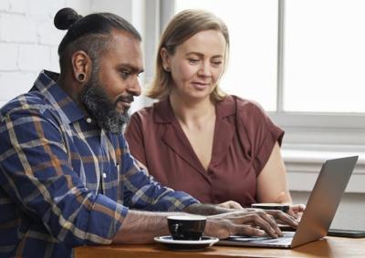 A stock photo of two people in an office environment looking at the same laptop screen. A male-presenting human has a man bun and a plaid shirt, while a female-presenting human wears a maroon top.