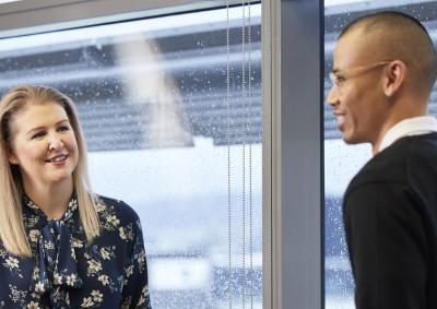 A man and a woman stand in front of a window, facing each other and having a conversation. They are smiling. 