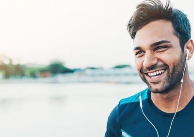 Picture of a male presenting human wearing headphones while outside. He has a smile on his face.