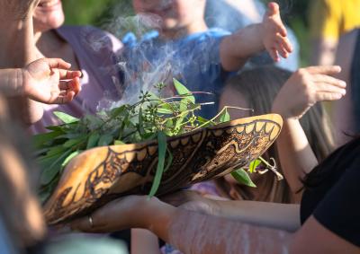 Image of a smoking ceremoney. A dish with leaves and smoke and hands pushing the smoke over people.