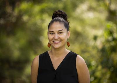 Photograph of a smiling Indigenous person with their hair tied up in a bun. They are wearing a sleeveless black top against a background of green leaves.