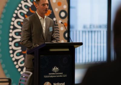 Image of Gary Rake standing at a lectern presenting to a crowd. Behind him is Aboriginal artwork. 