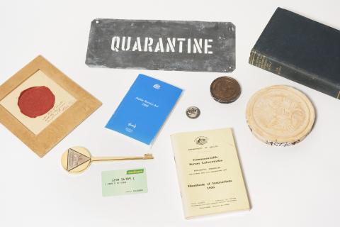 A group of 10 objects. Sign at the back includes a metal ‘Quarantine’ sign. At the front is a large gold key and a Medicare card.