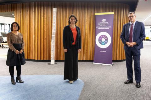 Three people in business attire stand in an office in front of a banner for the APS Mental Health Capability Framework Pilot Site.. 