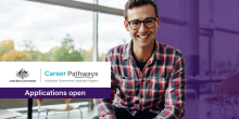 A young man wearing glasses and a checked shirt sits in an office smiling at the camera. Text reads: “Australian Government Career Pathways. Applications open.”